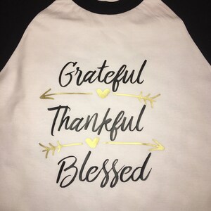 May include: A white baseball tee shirt with black sleeves. The shirt has a gold foil design with the words "Grateful", "Thankful", and "Blessed" with arrows and hearts.