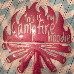 May include: A pink glitter iron-on design with the text "This is my campfire hoodie" on a white background.
