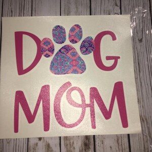 May include: A white decal featuring the text "DOG MOM" in pink glitter lettering. The letter "O" in "DOG" is replaced with a paw print design in pink, blue, and purple paisley patterns.