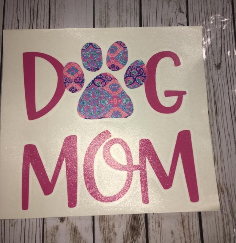 May include: A white decal featuring the text "DOG MOM" in pink glitter lettering. The letter "O" in "DOG" is replaced with a paw print design in pink, blue, and purple paisley patterns.
