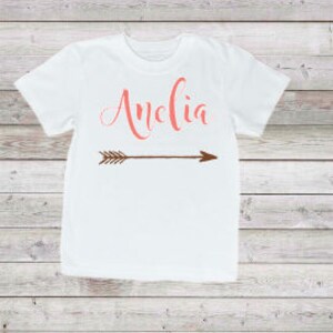 May include: White t-shirt with the name "Amelia" written in pink script and a brown arrow underneath.