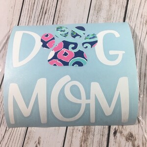 May include: A light blue vinyl decal with the words "DOG MOM" in white. A paw print graphic in the centre features a pattern of pink, blue, and white. The decal is designed for application on various surfaces.
