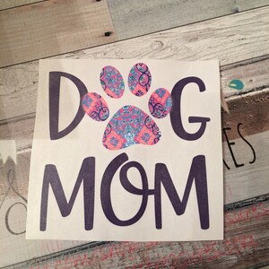 May include: A white vinyl decal featuring the text "DOG MOM" in a dark purple font. The letter "O" in "DOG" is replaced with a paw print design in pink, blue, and white paisley patterns. Set against a weathered wood backdrop.
