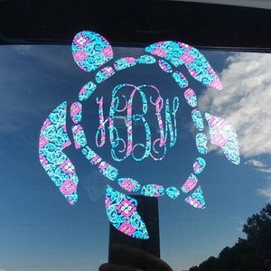 May include: A blue and pink patterned sea turtle decal with a monogram in the center. The decal is on a car window.