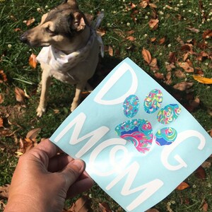 May include: A light blue sticker with the words "DOG MOM" in white, featuring a paw print design with colourful patterns. The sticker is held in a hand, with a dog in the background.