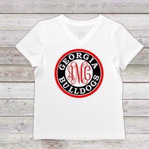 May include: White V-neck t-shirt with a red and black circle graphic that says "Georgia Bulldogs" with a monogram in the center.