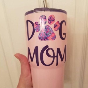 May include: A pink tumbler with the words "Dog Mom" in dark purple lettering. A paw print design in shades of pink, blue, and purple is above the word "Mom". The tumbler has a silver straw and a dark purple lid.