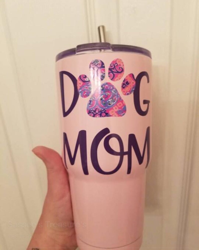 May include: A pink tumbler with the words "Dog Mom" in dark purple lettering. A paw print design in shades of pink, blue, and purple is above the word "Mom". The tumbler has a silver straw and a dark purple lid.