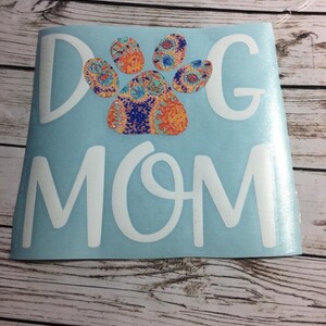 May include: A light blue decal with the words "DOG MOM" in white, featuring a colourful paw print design. The paw print has orange, blue, and yellow patterns. The decal is on a wooden surface.