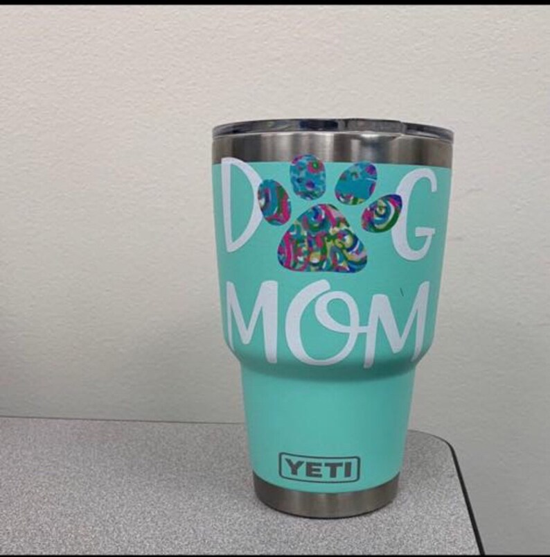 May include: A teal Yeti tumbler with the words "Dog Mom" in white lettering. A colourful paw print design is in the centre. The tumbler has a silver rim and the Yeti logo at the bottom.