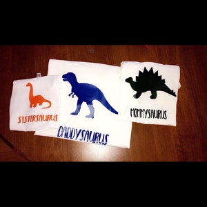 May include: Three white t-shirts with dinosaur silhouettes and the words "Sistersaurus", "Daddysaurus", and "Mommysaurus" printed on them.