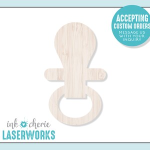 May include: A wooden cutout of a baby rattle in the shape of a ring and a teardrop. The cutout is a light brown color and has a wood grain texture. The text "ACCEPTING CUSTOM ORDERS MESSAGE US WITH YOUR INQUIRY" is in a blue circle in the upper right corner of the image. The text "ink chérie LASERWORKS" is in the lower left corner of the image.