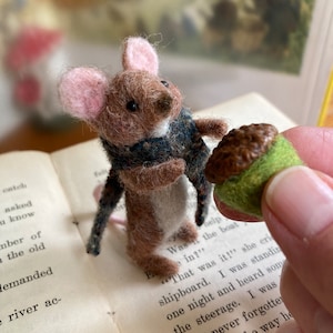 May include: A small brown mouse wearing a blue and white scarf holds a brown acorn with a green cap. The mouse is sitting on a book with text about a shipboard.