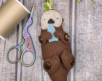 May include: A handmade brown felt otter ornament holding a blue felt fish, with button accents. A spool of beige thread and rainbow scissors are next to the otter. A green leaf sprig is attached to the otter's head.