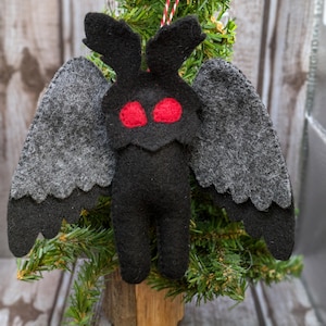 May include: A felt ornament shaped like a moth with large gray wings and red eyes. The ornament is hanging from a red and white string on a small artificial Christmas tree.
