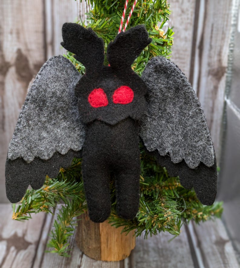 May include: A felt ornament shaped like a moth with large gray wings and red eyes. The ornament is hanging from a red and white string on a small artificial Christmas tree.