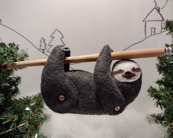 May include: A handmade felt sloth toy in dark gray, hanging from a wooden dowel. The sloth has a white face with brown accents and button details. The background features a simple drawing of a house and trees.