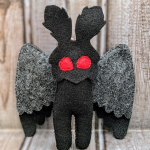 May include: A black felt plush toy shaped like a moth with large red eyes and gray wings.