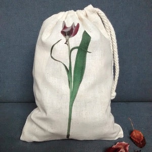 Puede incluir: Una bolsa de algodón blanco con cordón de ajuste con un estampado floral de un solo tulipán con una flor de color burdeos y hojas verdes.