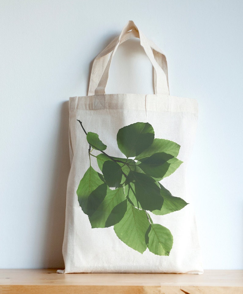 K&ouml;nnte beinhalten: Eine wei&szlig;e Stofftasche mit einem gr&uuml;nen Blattdesign, das auf der Vorderseite aufgedruckt ist. Die Bl&auml;tter sind in einem ast&auml;hnlichen Muster angeordnet.