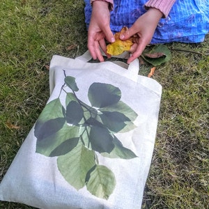 K&ouml;nnte beinhalten: Eine wei&szlig;e Stofftasche mit einem gr&uuml;nen Blattdesign. Die Tasche hat zwei Henkel und liegt auf einem Bett aus gr&uuml;nem Gras.