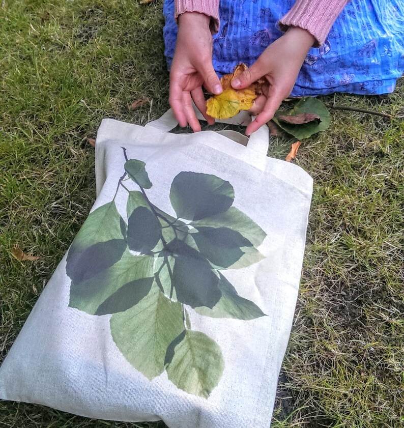 K&ouml;nnte beinhalten: Eine wei&szlig;e Stofftasche mit einem gr&uuml;nen Blattdesign. Die Tasche hat zwei Henkel und liegt auf einem Bett aus gr&uuml;nem Gras.