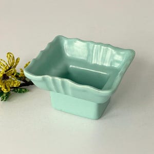 May include: A light mint green ceramic square bowl with a pedestal base and a wavy, scalloped rim. Beside the bowl is a small decorative sprig of yellow flowers with green leaves, crafted from tiny beads on thin wire stems.