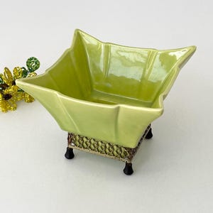 May include: A light green ceramic bowl with a starburst shape, set on a decorative metal stand with black feet. The bowl has a glossy finish. A beaded floral accent is to the left.