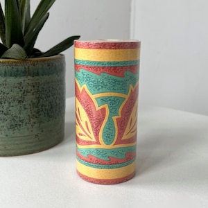 May include: A cylindrical decorative container with a colorful floral design. The container features bands of yellow, teal, and red, with a repeating pattern of stylized flowers. The container is standing upright on a white surface.