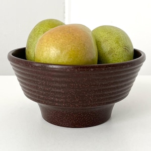May include: A dark brown ceramic bowl with a textured, ridged exterior, holding three pears. The pears vary in color, with some green and some with a yellow-brown hue. The bowl sits on a white surface.