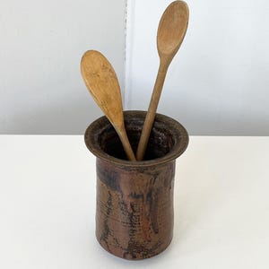 May include: A cylindrical brown ceramic crock with a wide rim and a dark, mottled glaze finish. Two light-colored wooden spoons are placed inside the vessel, with their handles extending upward from the opening of the container.