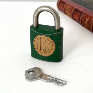 May include: A green padlock with a silver shackle and a gold-colored circular emblem. A silver key rests nearby. The lock is next to a vintage book with a red cover.