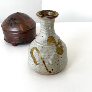 May include: A small, speckled ceramic vase with a narrow neck and a rounded body, adorned with brown floral designs. Beside it is a small, round, lidded container with a dark brown finish. Both items are on a white surface.