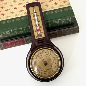 May include: A vintage brown Airguide barometer and thermometer. The thermometer displays a temperature reading, and the barometer has a dial indicating weather conditions. The device is set against a backdrop of antique books.