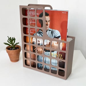 May include: A taupe magazine rack with a handle, holding a magazine with a man on the cover. The rack has a grid-like design with square cutouts. A small potted plant sits to the left of the rack on a white surface.