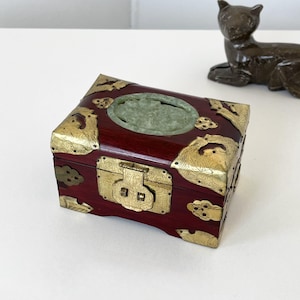 May include: A small, rectangular wooden box with a dark red finish and ornate brass hardware. The box features a jade-colored oval inset on the lid and decorative cloud-shaped accents. A small bronze cat figurine is in the background.