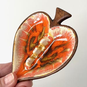 May include: A leaf-shaped ashtray with a vibrant orange and cream interior, framed by a dark brown border. The ashtray features a raised section in the center and a small, pointed handle. The design is reminiscent of mid-century modern decor.