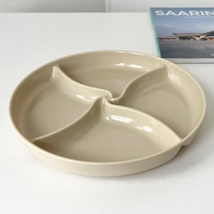 May include: A beige, round divided serving dish with four sections. The dish is made of a smooth material and is designed for serving snacks or appetizers. A book titled "SAARINEN" is in the background.