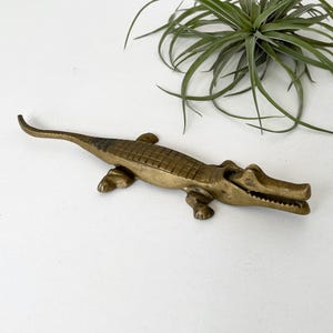 May include: A brass alligator figurine featuring a textured, scaled back, four legs, and an open mouth with visible teeth. The metal surface has a muted gold tone with darker patina accents along the spine and tail, capturing a realistic form.