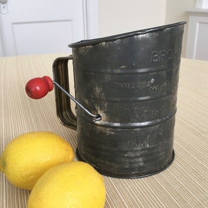 May include: An antique black metal flour sifter with a red wooden handle and a wire agitator. The sifter is labeled "BROWN" and "THREE CUPS MEASURE". Two bright yellow lemons sit beside the sifter on a striped surface.