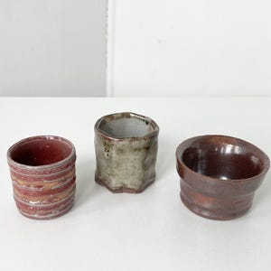May include: Three handmade ceramic vessels in varying shapes and colors. The leftmost cup is red with horizontal bands. The center cup is gray with a textured surface. The rightmost bowl is brown with a slanted rim.