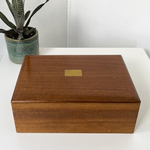 May include: A rectangular wooden box with a smooth, polished finish and a prominent wood grain pattern. The lid features a centered, small, rectangular brass-colored metal plate. The box has a hinged construction and a deep, warm brown tone.