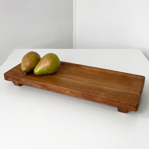 May include: A rectangular wooden serving tray with two pears. The tray is made of a warm-toned wood and has small feet. The pears are green and brown, and the tray is empty.