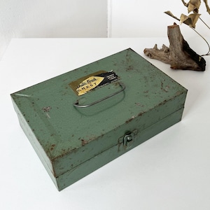 May include: A rectangular, sage green metal storage chest with a weathered, rusted finish. It features a metal carrying handle on the lid, a front latch, and a yellow and black label that reads Multi-Torch CHEST TURNER BRASS WORKS SYCAMORE ILLINOIS.