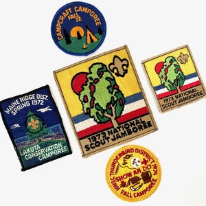 May include: A collection of vintage embroidered patches. The patches feature text such as "Campcraft Camporee Fall '72", "1973 National Scout Jamboree", and "Maine Ridge Dist. Spring 1972". The patches are in various shapes and sizes, with colorful designs.
