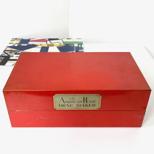May include: A rectangular, red box with a shiny finish. A gold-colored label on the front reads "The American Home Menu Maker." A colorful, patterned book is partially visible in the background.