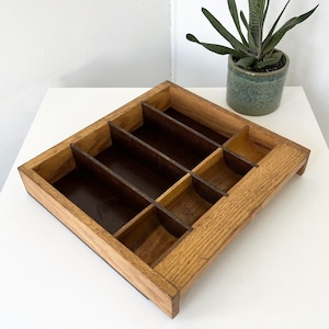 May include: A rectangular wooden organizer tray featuring a light brown outer frame and darker brown interior dividers. The tray is divided into four long vertical compartments and two smaller square compartments, creating a structured storage space.