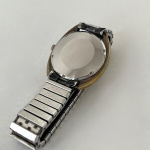 May include: A wristwatch with a round, silver-toned metal case back and a gold-toned bezel. The watch features a silver-toned, expandable metal link band with the brand name Speidel and patent numbers engraved on the individual links.