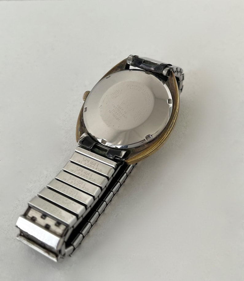 May include: A wristwatch with a round, silver-toned metal case back and a gold-toned bezel. The watch features a silver-toned, expandable metal link band with the brand name Speidel and patent numbers engraved on the individual links.