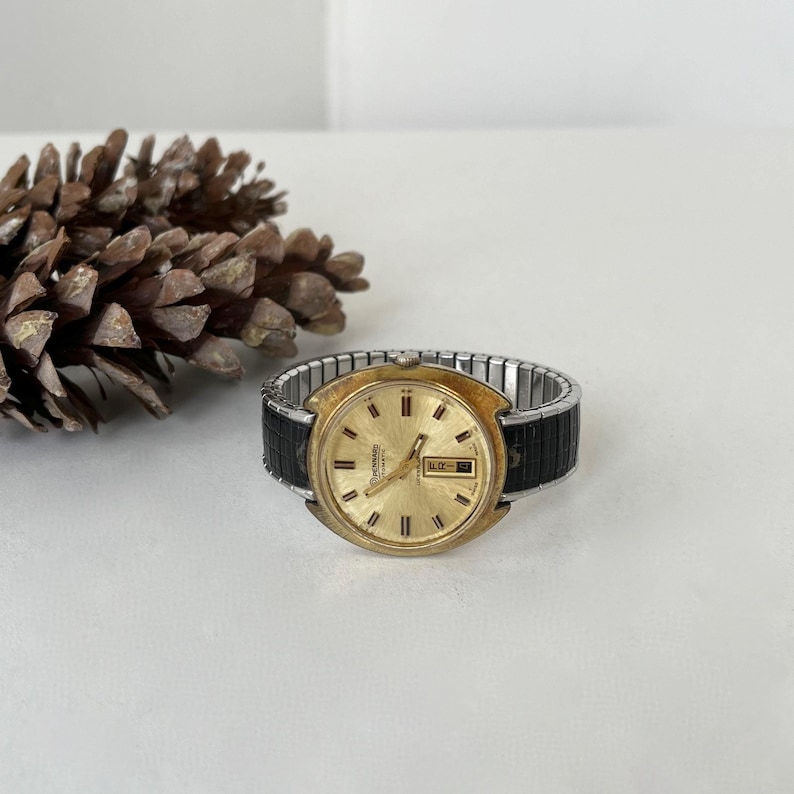 May include: A vintage Pennard automatic wristwatch with a round gold-tone dial, gold-tone hour markers, and a day-date window at the three o'clock position. The watch features a two-tone expansion band with black textured links and silver-tone metal.